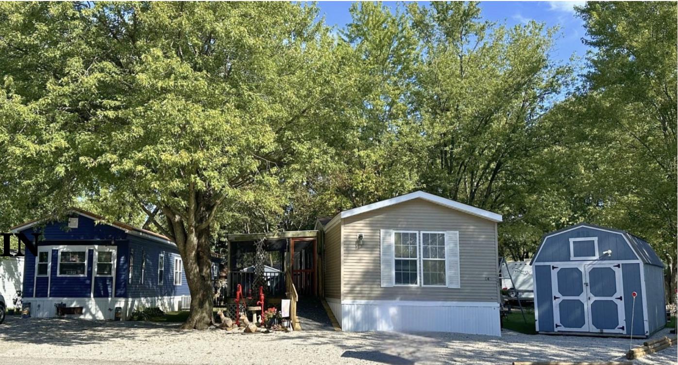 Two manufactured homes facing a gravel street at Thunder Hill: a cream home with white trim and a screened porch, a blue home to its left, and a blue-and-white storage shed to the right, under mature trees.