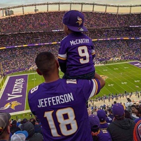 A man in a Vikings jersey at a football game, with a child on his shoulders, both seen from behind.