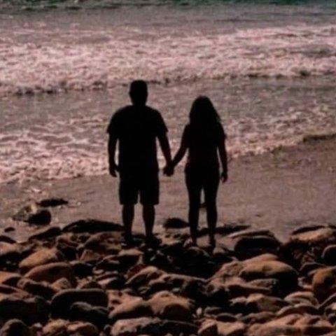 Two people holding hands on a rocky beach, in silhouette.
